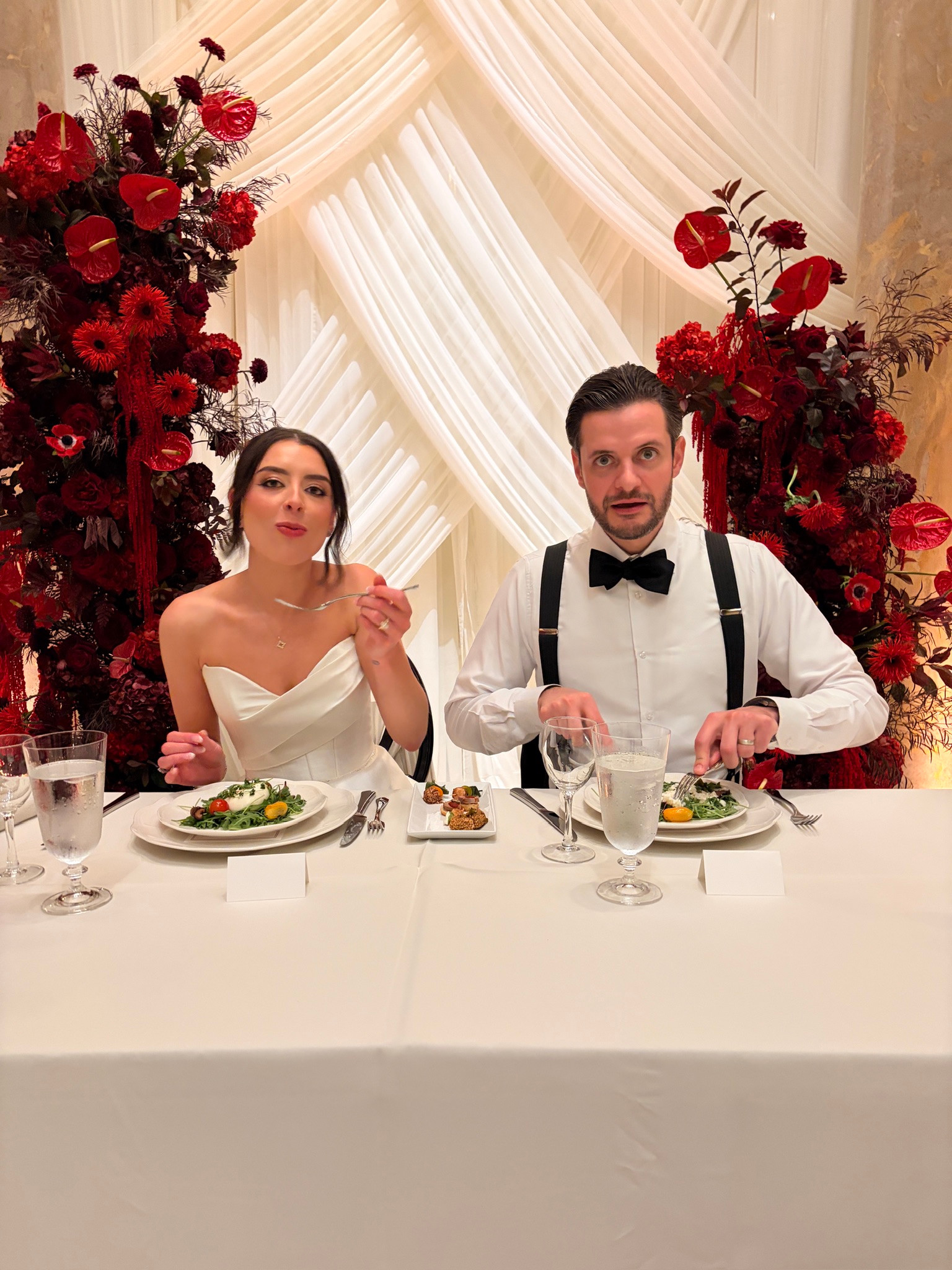 Kayla and Chris at their wedding reception, surrounded by deep red flowers and ivory drapery.